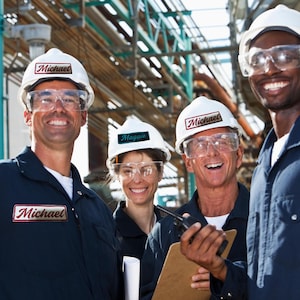 May include: Four workers wearing navy blue coveralls, white hard hats with name tags, and safety glasses. The name "Michael" is visible on two hats and a patch. One worker holds a clipboard and radio. The background shows industrial piping.
