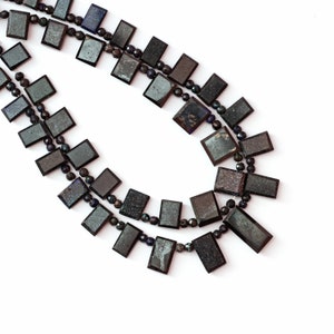 Puede incluir: Un collar de doble cadena hecho de cuentas de piedras preciosas negras e iridiscentes. Las cuentas son rectangulares y tienen un acabado brillante.