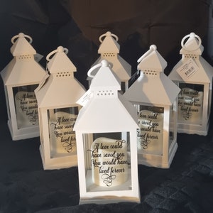 May include: A set of six white candle lanterns with glass panels. Each lantern features a white candle with a black inscription that reads "If love could have saved you, you would have lived forever".