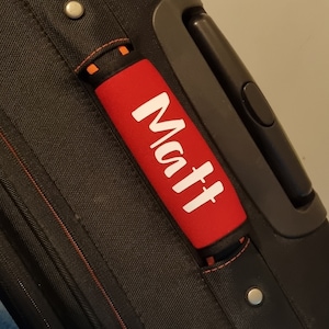 May include: Black suitcase with a red handle wrap. The handle wrap has the name "Matt" printed in white. The suitcase has a black handle and silver rivets.