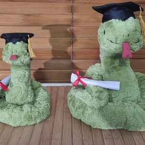 May include: Two plush green snakes wearing graduation caps with gold tassels. One snake holds a diploma tied with a red ribbon. The other snake also holds a diploma. The snakes are on a wooden surface.