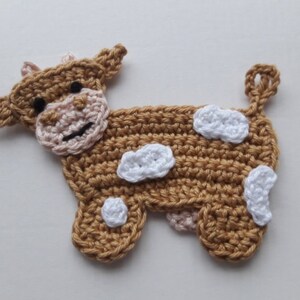 May include: A crocheted brown cow with white spots. The cow has a pink nose and black eyes.