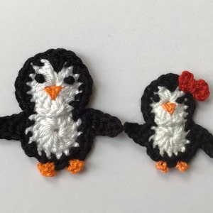May include: Two crocheted penguin ornaments, one larger than the other. The larger penguin is black and white with orange feet. The smaller penguin is black and white with a red bow and orange feet.