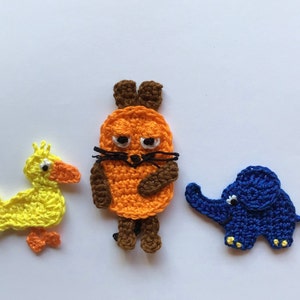 Pode incluir: Três figuras de animais de crochê: um pato amarelo, um gato laranja e um elefante azul. Os animais têm olhos brancos e são feitos de fio.