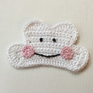 Könnte beinhalten: Weiße gehäkelte Wolke mit einem lächelnden Gesicht, schwarzen Augen und rosa Wangen.