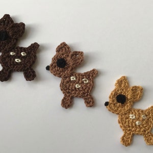 Könnte beinhalten: Drei gehäkelte Hirsch-Ornamente in Braun, Beige und Gold. Jeder Hirsch hat weiße Flecken und schwarze Augen.