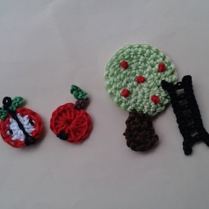 Op de afbeelding: Gebreide appelversieringen in rood en groen met een zwarte ladder en een groene boom met rode appels.