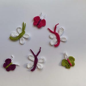 Peut inclure: Six papillons en crochet de différentes couleurs, notamment vert, blanc, rose et violet. Les papillons sont disposés sur un fond blanc.