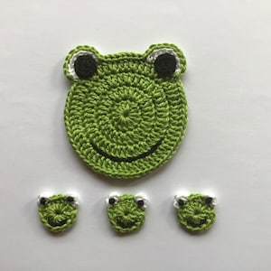 Könnte beinhalten: Grüne gehäkelte Frosch-Applikation mit einem lächelnden Gesicht und schwarzen Augen. Drei kleinere grüne gehäkelte Frosch-Applikationen mit schwarzen Augen sind ebenfalls enthalten.