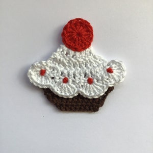 Könnte beinhalten: Eine gehäkelte Cupcake-Applikation mit weißem Frosting, braunem Boden und roter Kirsche oben drauf.