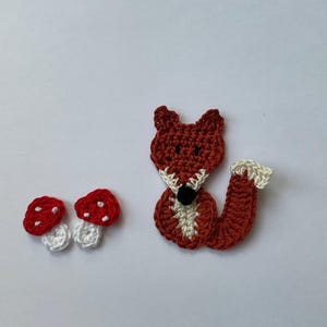 Könnte beinhalten: Ein gehäkelter Fuchs mit einer weißen Brust und einem roten Schwanz, der neben zwei roten und weißen gehäkelten Pilzen sitzt.