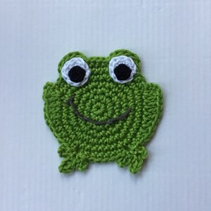 May include: Green crocheted frog applique with black eyes and a white outline. The frog has a smiling mouth.
