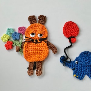 Può includere: Un gatto arancione all'uncinetto che tiene un mazzo di fiori colorati, un elefante blu all'uncinetto che tiene un palloncino rosso e un palloncino rosso all'uncinetto.