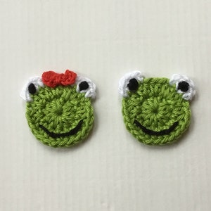 Könnte beinhalten: Zwei grüne gehäkelte Froschgesichter mit schwarzen Augen, weißen Akzenten und schwarzen lächelnden Mündern. Ein Frosch hat eine rote Schleife auf dem Kopf.