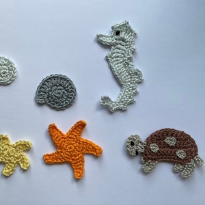 Puede incluir: Una colección de criaturas marinas de ganchillo, incluyendo un caballito de mar, estrellas de mar, una tortuga y conchas marinas. El caballito de mar es verde claro, las estrellas de mar son naranjas y amarillas, y la tortuga es marrón con manchas blancas. Las conchas son grises y verde claro.