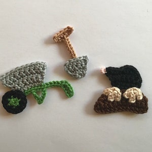 Könnte beinhalten: Gehäkeltes Garten-Set mit einer grauen Schubkarre mit grünem Griff und schwarzem Rad, einer braunen und grauen Schaufel und einem schwarzen Maulwurf mit beigefarbenen Pfoten. Ideal für Bastelarbeiten.