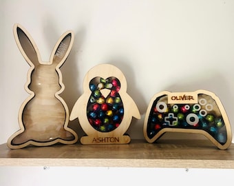 May include: Three wooden shadow box banks with clear acrylic fronts. One is shaped like a rabbit with the name Maddie, one like a penguin with the name Ashton, and one like a game controller with the name Oliver. The penguin and controller contain colorful foil-wrapped candies.