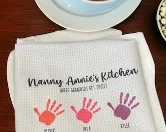 May include: White waffle-weave kitchen towel with the text "Nanny Annie's Kitchen Where Grandkids Get Spoilt." Features three colorful handprints with the names Oliver, Ayla, and Rylee. A bowl of strawberries sits above.