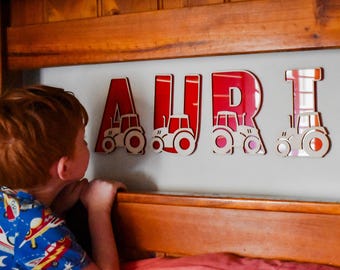 May include: Red and white wooden letters spelling the name AURI, each featuring a tractor design. The letters are mounted on a light gray wall. A young child is looking at the letters.