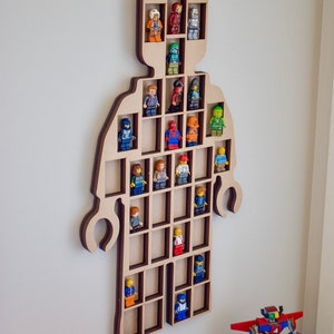 May include: A wall-mounted wooden display shelf shaped like a large toy figure with multiple rectangular compartments. The light-colored wood frame holds various small plastic character figurines in individual slots, showcasing a collection of figures.