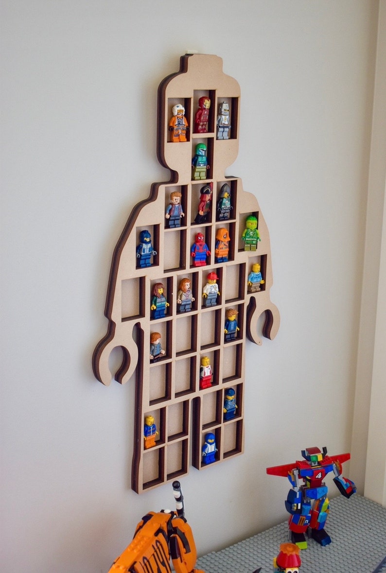 May include: A wall-mounted wooden display shelf shaped like a large toy figure with multiple rectangular compartments. The light-colored wood frame holds various small plastic character figurines in individual slots, showcasing a collection of figures.