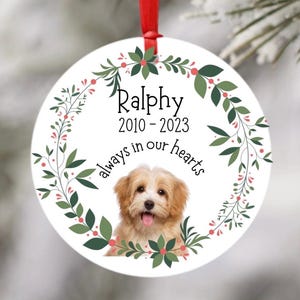 May include: White round Christmas bauble with a green and red floral wreath, a photo of a small dog, and the text "Ralphy 2010 - 2023 always in our hearts".