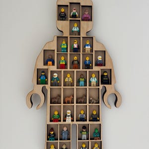 May include: A light-colored wooden wall-mounted display shelf shaped like a toy figure. The shelf features multiple rectangular compartments containing various small plastic toy figures, including people and animals, arranged in rows within the frame.
