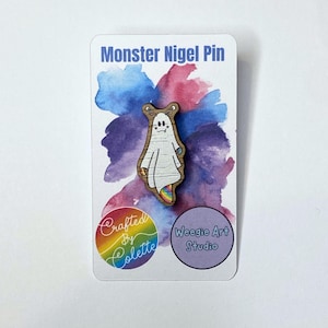 May include: Wooden pin featuring a cartoon ghost with a rainbow tail. The ghost has a surprised expression with large eyes and a small mouth. The pin is on a white card with the text "Monster Nigel Pin" and "Weegie Art Studio".