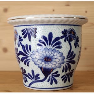 May include: A ceramic planter with a white and blue floral design. The planter has a light blue background with a repeating pattern of small dots. Large blue flowers and leaves are painted around the planter. The rim is white with a gold trim.