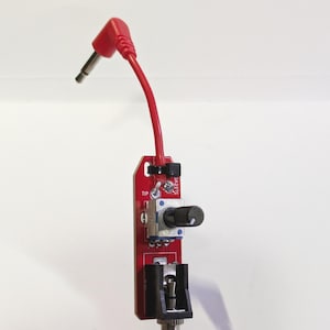 Könnte beinhalten: Eine rote Platine mit einem schwarzen Knopf und einem rot-schwarzen Kabel mit einem 6,35 mm Stecker. Auf der Platine sind die Worte "TIP" und "SLEEVE" aufgedruckt.
