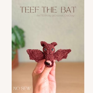 May include: A small, dark brown crocheted bat plush toy with two black plastic eyes and two tiny white fangs. The text TEEF THE BAT, PATTERN by @Crochet_CroChey, and NO SEW are visible. The bat has pointed ears and outstretched wings.