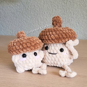 Op de afbeelding: Twee gehaakte eikel knuffels met bruine dopjes en witte lichamen. De knuffels hebben zwarte ogen en glimlachende monden. De knuffels zitten op een houten oppervlak. De tekst "@crochet_CroChey" is zichtbaar op de achtergrond.