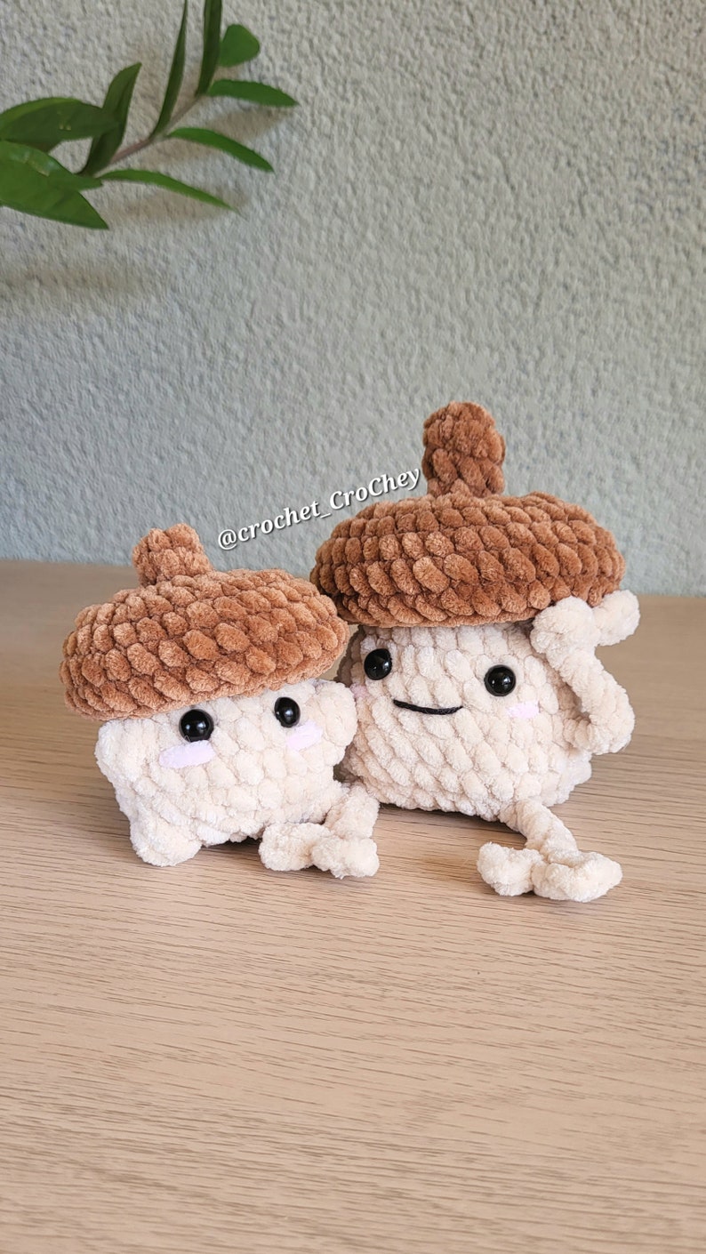 Op de afbeelding: Twee gehaakte eikel knuffels met bruine dopjes en witte lichamen. De knuffels hebben zwarte ogen en glimlachende monden. De knuffels zitten op een houten oppervlak. De tekst "@crochet_CroChey" is zichtbaar op de achtergrond.
