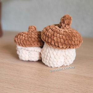 Op de afbeelding: Twee gebreide eikelvormige containers, een groter dan de andere. De grotere eikel is bruin met een witte bovenkant, en de kleinere eikel is bruin met een witte bovenkant. De eikels staan op een lichtbruin houten oppervlak. @crochet_Crochey staat op het oppervlak.