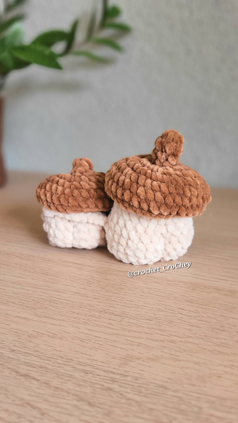 Op de afbeelding: Twee gebreide eikelvormige containers, een groter dan de andere. De grotere eikel is bruin met een witte bovenkant, en de kleinere eikel is bruin met een witte bovenkant. De eikels staan op een lichtbruin houten oppervlak. @crochet_Crochey staat op het oppervlak.