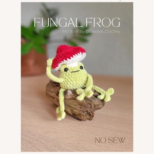 May include: A crocheted frog wearing a red and white mushroom hat, sitting on a piece of driftwood. The frog is light green with black eyes and is labeled "Fungal Frog" with the text "PATTERN by @Crochet_Crohey" and "NO SEW".