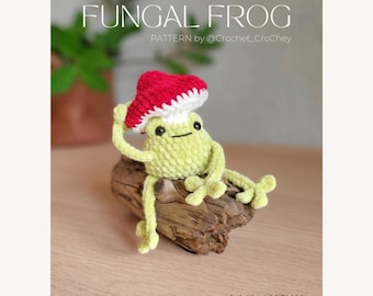 May include: A crocheted frog wearing a red and white mushroom hat, sitting on a piece of driftwood. The frog is light green with black eyes and is labeled "Fungal Frog" with the text "PATTERN by @Crochet_Crohey" and "NO SEW".