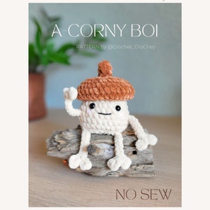 Pode incluir: Um brinquedo de crochê em forma de bolota, com uma tampa marrom e um corpo branco, sentado em um pedaço de madeira flutuante. O brinquedo tem um rosto sorridente e dois braços levantados no ar. O texto "A-CORNY BOI" e "NO SEW" são visíveis na imagem. PATTERN by @Crochet_Crochey