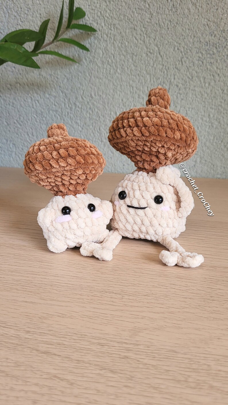 Op de afbeelding: Twee gehaakte paddenstoel knuffels met bruine viltmutsen en witte lichamen. De knuffels hebben geborduurde ogen en glimlachen. De knuffels zitten op een houten oppervlak.