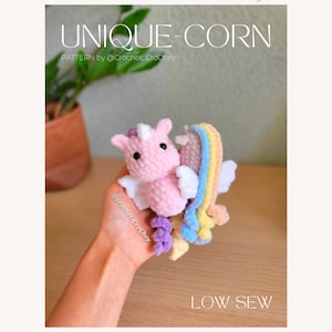 May include: A pink crocheted unicorn with a rainbow tail and white wings. The unicorn is being held in a hand. The text "UNIQUE-CORN" and "LOW-SEW" is visible in the image.