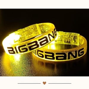 May include: Two translucent yellow plastic bracelets that emit a bright glow. Each circular band features the word BIGBANG printed in bold black capital letters. The material has a textured, glittery appearance and a visible clasp mechanism.