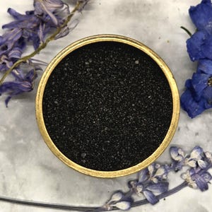 May include: A round, gold-colored container filled with black granules. The container is set against a backdrop of dried purple and blue flowers. The granules fill the container to the top.