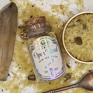 May include: A clear glass jar with a cork stopper filled with light yellow sea salt. The label reads Mystic Moon Apothecary Open Road No More Detours Abre Camino Natural Sea Salt. A gold key charm hangs from the neck, next to a crystal and a bowl of salt.