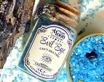 May include: A clear glass jar filled with blue sea salt, sealed with a cork and twine. The label reads Mystic Moon Apothecary, Evil Eye, Can't Hex This, and Cedarwood Natural Sea Salt, featuring an eye illustration and moon phases.