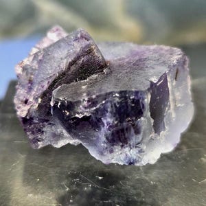May include: Close-up of a fluorite crystal with a mix of purple and clear sections. The crystal has a rough, textured surface and a geometric shape. The lighting highlights the different shades of purple and the crystal's natural formations.