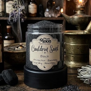 May include: A jar of black cauldron sand from Mystic Moon Apothecary. The label reads "Cauldron Sand Black" with the words "Protection, Banishing, Warding, Absorbing". The jar sits on a black base, with other apothecary items in the background.