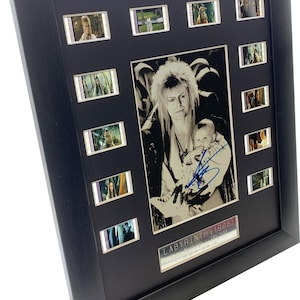 Pode incluir: Uma moldura preta com uma fotografia central a preto e branco de uma pessoa com cabelo espetado a segurar um bebé, assinada com tinta azul. A moldura inclui doze pequenas tiras de filme à volta da foto e uma placa LABYRINTH (1986).