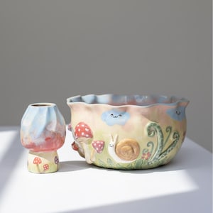 Puede incluir: Un conjunto de maceta y jarrón de cerámica caprichosos. El jarrón tiene forma de hongo con un diseño azul, rojo y amarillo. La maceta presenta un borde ondulado, un caracol, setas y detalles de nubes. La paleta de colores incluye tonos pastel de amarillo, rosa y azul.