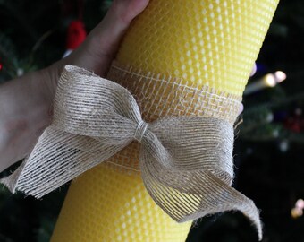 May include: A large, cylindrical yellow beeswax candle with a textured surface, wrapped with a burlap ribbon tied in a bow. The candle is approximately 8 inches tall and is a decorative item.