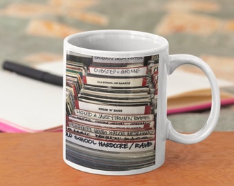 May include: A white ceramic mug with a colorful image of a stack of vinyl records. The records are labeled with different music genres, including "Dubstep + Grime", "Drum 'n' Bass", "Liquid & Jazzy Drum'n' Bass", and "Old School Hardcore/Rave".